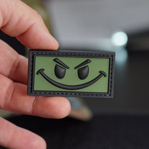 May include: A black and green embroidered patch with a smiling face with angry eyes. The patch has a black stitched border.