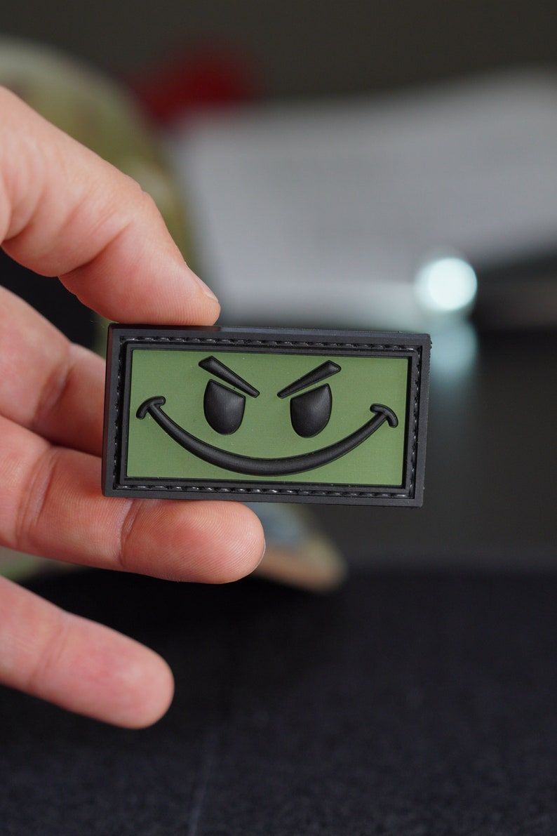 May include: A black and green embroidered patch with a smiling face with angry eyes. The patch has a black stitched border.