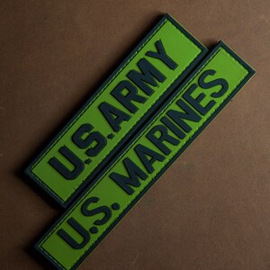 Può includere: Due toppe verdi con cuciture nere e testo nero. La toppa superiore dice "U.S. ARMY" e la toppa inferiore dice "U.S. MARINES".
