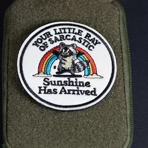 Pode incluir: Um emblema bordado circular com fundo branco e borda preta. Apresenta um guaxinim cinzento sob um arco-íris colorido. O texto diz: YOUR LITTLE RAY OF SARCASTIC Sunshine Has Arrived, centralizado em letras pretas sobre o emblema.