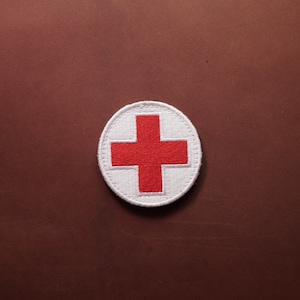 Peut inclure: Un écusson rond avec une croix rouge sur fond blanc. Le patch est brodé avec un motif détaillé et possède une bordure cousue. Le patch est un symbole d'aide médicale et de services d'urgence.