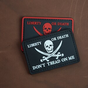 Puede incluir: Dos parches negros y blancos con un diseño de calavera y huesos cruzados. El parche superior dice "Libertad o muerte" en letras rojas. El parche inferior dice "Libertad o muerte" en letras blancas y "No pises sobre mí" en letras blancas.