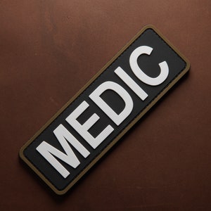 Pu&ograve; includere: Un cerotto nero e bianco con un bordo marrone. Il cerotto dice "MEDIC" in lettere bianche.
