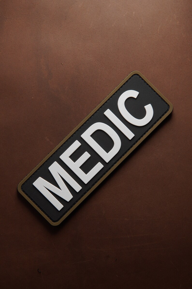 Pu&ograve; includere: Un cerotto nero e bianco con un bordo marrone. Il cerotto dice "MEDIC" in lettere bianche.