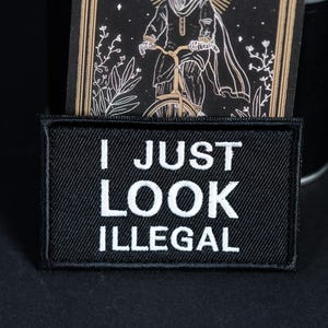 Op de afbeelding: Een rechthoekige zwarte geborduurde patch met de witte tekst I JUST LOOK ILLEGAL in een vetgedrukt schreefloos lettertype. De patch heeft een gestikte rand en is geplaatst voor een zwarte kaart met een gouden lijntekening van een persoon op een fiets.