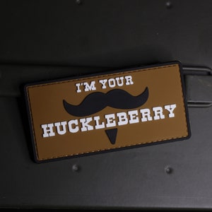 Może przedstawiać: Brązowa łatka z czarnymi szwami i białym tekstem, który brzmi "I'm your Huckleberry" z czarnym wzorem wąsów.