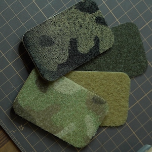 Puede incluir: Cuatro muestras de tela de camuflaje en diferentes colores: verde, marrón y negro. Las muestras están dispuestas en una pila sobre una superficie de rejilla negra.