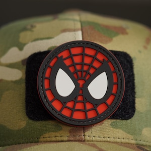 May include: A round, red, black, and white Spider-Man patch. The patch features the character's iconic mask design with a red web pattern, black outline, and white eye shapes. The patch is attached to a camouflage hat.