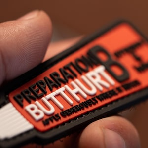 May include: A rectangular patch with the words "PREPARATION BUTTHURT" in white and black lettering on a red background. The text "APPLY GENEROUSLY WHERE IT HURTS" is also visible. The patch is black around the edges.