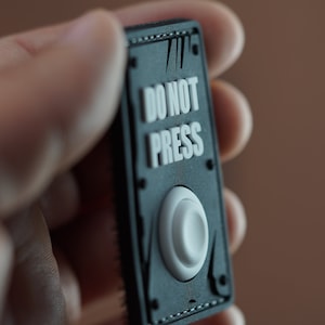 May include: A black and white button with the text "DO NOT PRESS" on it. The button is surrounded by a black frame with a textured edge.