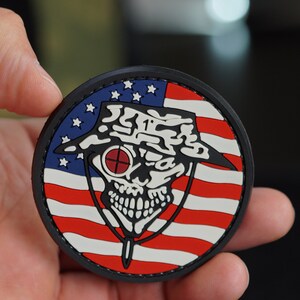 Könnte beinhalten: Ein schwarzer und weißer Schädel, der einen Tarnhut mit einem roten Fadenkreuz über dem Auge trägt. Der Schädel ist auf eine amerikanische Flagge mit roten und weißen Streifen und blauen Sternen gelegt.