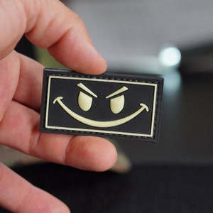 May include: A black patch with a yellow smiling face with raised eyebrows. The patch has a black stitched border.