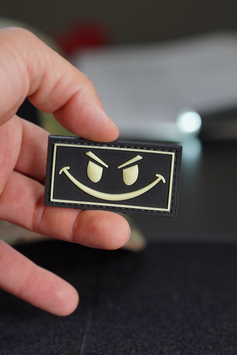 May include: A black patch with a yellow smiling face with raised eyebrows. The patch has a black stitched border.