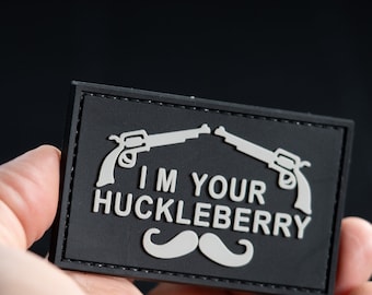 Może przedstawiać: Czarna prostokątna naszywka z napisem "I'M YOUR HUCKLEBERRY" i stylizowanymi wąsami. Dwa srebrne rewolwery umieszczone są nad tekstem. Naszywka trzymana jest w dłoni.
