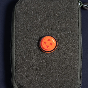 May include: A circular orange charm featuring four red stars, resembling a Dragon Ball, attached to the center of a rectangular olive green zippered pouch with a textured fabric surface and a dark zipper pull visible at the top corner.