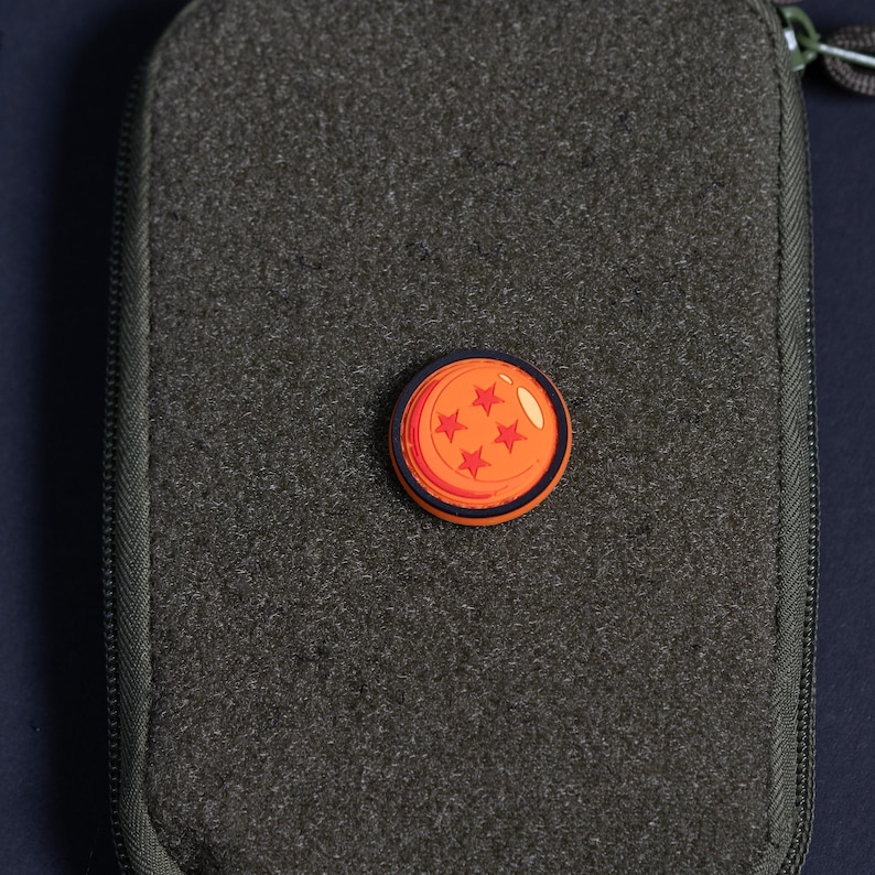 May include: A circular orange charm featuring four red stars, resembling a Dragon Ball, attached to the center of a rectangular olive green zippered pouch with a textured fabric surface and a dark zipper pull visible at the top corner.