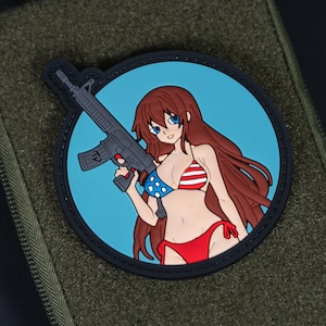 Könnte beinhalten: Runder Aufnäher mit einer Anime-Frau im Bikini, die ein Gewehr hält. Das Bikini-Oberteil ist mit Sternen und Streifen verziert, vor einem hellblauen Hintergrund. Der Aufnäher hat einen schwarzen Rand und befindet sich auf einem grünen Stoff.