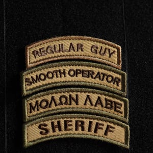 Op de afbeelding: Vier geborduurde patches in tan en bruin met zwarte stiksels. De patches zijn verticaal gerangschikt, met de bovenste patch met de tekst "REGULAR GUY", de tweede patch met de tekst "SMOOTH OPERATOR", de derde patch met de tekst "ΜΟΛΩΝ ΛΑΒΕ" en de onderste patch met de tekst "SHERIFF".