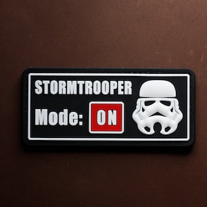 Peut inclure: Patch noir et blanc avec un casque de Stormtrooper et le texte "Stormtrooper Mode: ON" en lettres blanches sur fond rouge.