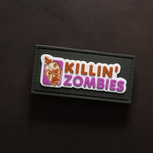 Può includere: Una toppa nera con cuciture bianche attorno ai bordi. La toppa ha una grafica rosa e arancione che dice "KILLIN' ZOMBIES" con un viso di zombie nell'angolo.