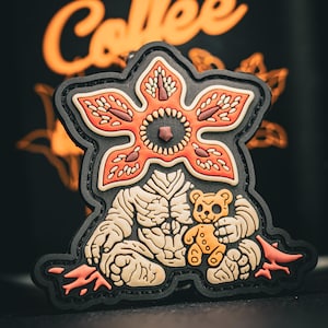 Może przedstawiać: Czarna naszywka przedstawiająca stworzenie podobne do Demogorgona trzymające misia. Stworzenie jest w odcieniach beżu, pomarańczu i czerwieni, z czarnym konturem. Słowo "Collée" jest pomarańczowym skryptem nad naszywką.