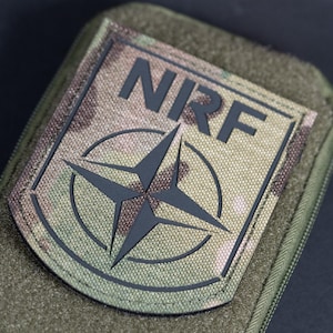 Puede incluir: Un parche táctico rectangular con un patrón de camuflaje en tonos verde, marrón y bronceado. El parche muestra el texto negro NRF sobre un emblema de estrella negra de cuatro puntas dentro de un borde circular, sobre un soporte de velcro.