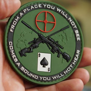 May include: Green and black embroidered patch with a world map, two rifles crossed, a playing card ace of spades, and the text "From a place you will not see comes a sound you will not hear."