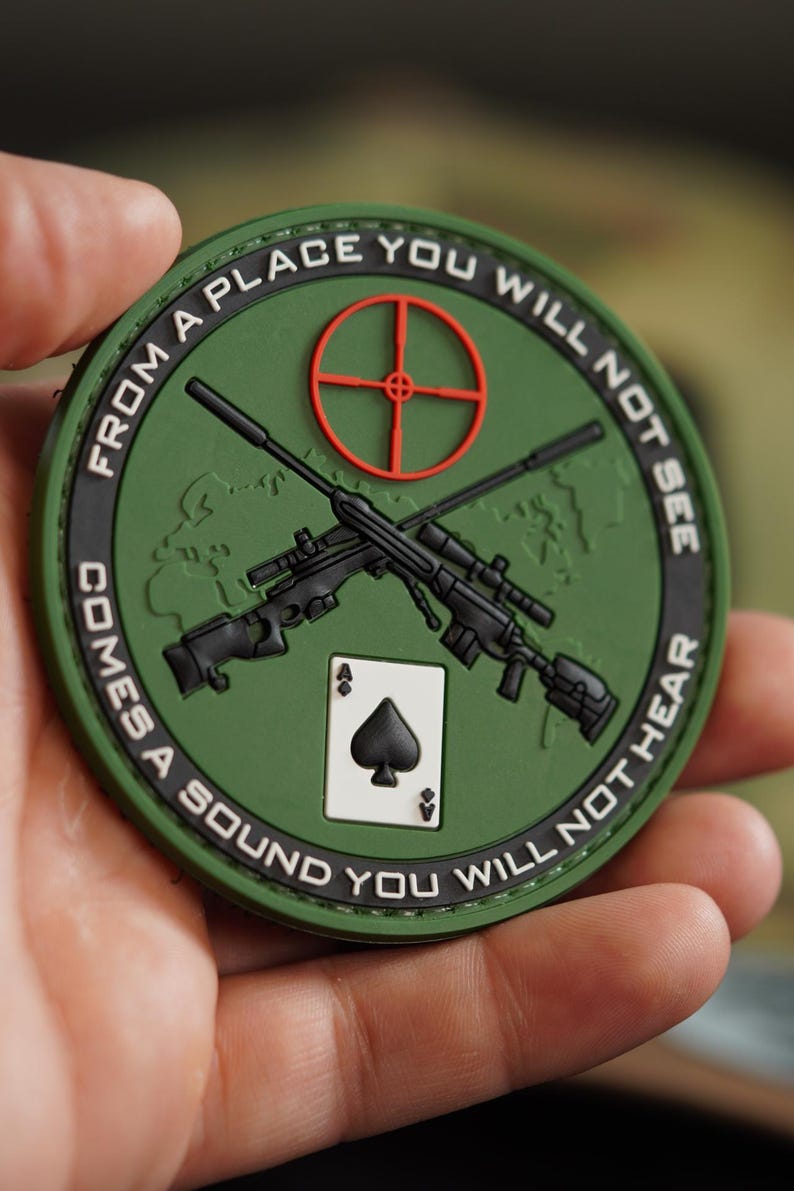 May include: Green and black embroidered patch with a world map, two rifles crossed, a playing card ace of spades, and the text "From a place you will not see comes a sound you will not hear."