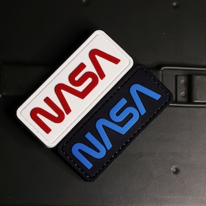 May include: Two NASA patches, one with red lettering on a white background and the other with blue lettering on a black background.