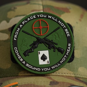 May include: A green, round patch with a white background and black text. The text reads "From a place you will not see comes a sound you will not hear". The patch features a red target, two crossed rifles, and a playing card with a black spade.