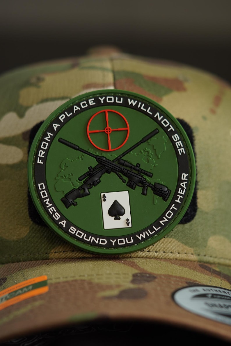 May include: A green, round patch with a white background and black text. The text reads "From a place you will not see comes a sound you will not hear". The patch features a red target, two crossed rifles, and a playing card with a black spade.