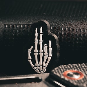 Könnte beinhalten: Ein schwarzer Gummibeschlag mit einem weißen Skelett-Hand-Design. Die Hand zeigt den Mittelfinger.