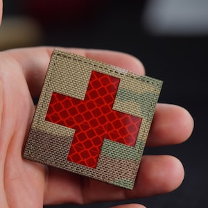 Könnte beinhalten: Ein kleiner, quadratischer, camouflagefarbener Patch mit einem roten reflektierenden Kreuz in der Mitte. Der Patch ist mit schwarzem Faden an den Rändern vernäht.