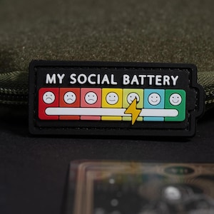 Può includere: Una toppa rettangolare nera con il testo bianco MY SOCIAL BATTERY sopra un indicatore di batteria orizzontale. L'indicatore è composto da sette segmenti colorati con faccine che vanno da triste a felice, con un fulmine giallo sul quinto.