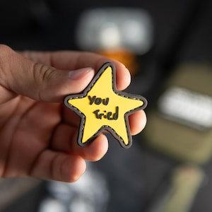 Op de afbeelding: Een stervormige patch met een geel midden en een donkergrijze rand. De tekst "You Tried" is in zwarte letters in het midden van de ster gedrukt. De patch heeft een gestikte randafwerking rondom de omtrek van de gele stervorm.