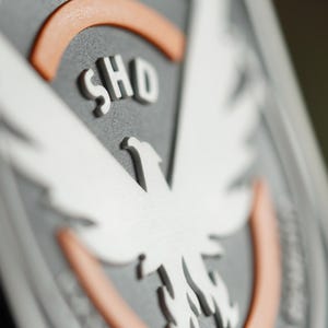 Könnte beinhalten: Nahaufnahme eines grauen Emblems mit einem weißen Adlerdesign und den Buchstaben "SHO". Das Emblem hat eine erhabene, strukturierte Oberfläche mit orangefarbenen und weißen Akzenten. Das Design ist detailliert und dreidimensional.