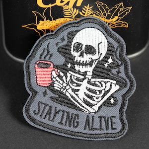 May include: An embroidered patch featuring a white skeleton holding a red striped mug with steam rising from it. The background is dark gray with black stitching details and small white stars. The text STAYING ALIVE is stitched in black at the bottom.