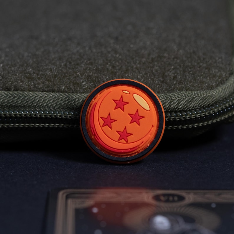 May include: A circular orange patch featuring a black border and four red star icons arranged in a diamond pattern, with a light yellow highlight on the upper right edge, designed as a decorative accessory for clothing or gear.