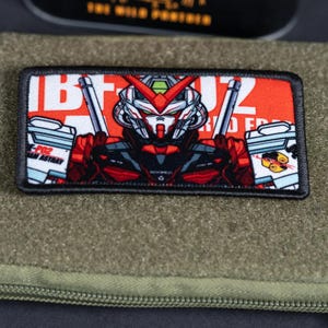 May include: A rectangular morale patch featuring a red and white graphic of a robotic character with a V-shaped crest. The design includes the text MBF-P02 and ASTRAY, with black embroidered borders, set against a textured olive green fabric background.