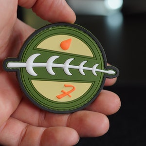 Könnte beinhalten: Ein runder grüner Patch mit einem weißen Knochenmuster, einem roten und orangefarbenen Hammer und Sichel und einem kleinen orangefarbenen Flüssigkeitstropfen. Der Patch hat einen gelben Hintergrund und einen grünen Rand.