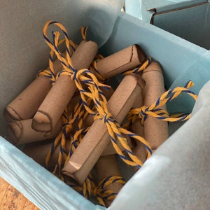 Könnte beinhalten: Eine Sammlung von aufgerollten Papierröhrchen, die mit gelber und blauer Schnur zusammengebunden sind. Die Röhrchen sind in einem korbartigen Behälter angeordnet.