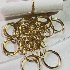 May include: A pile of gold-colored jump rings, also known as key rings, are scattered on a white surface. The rings are small and circular, and they are all the same size.