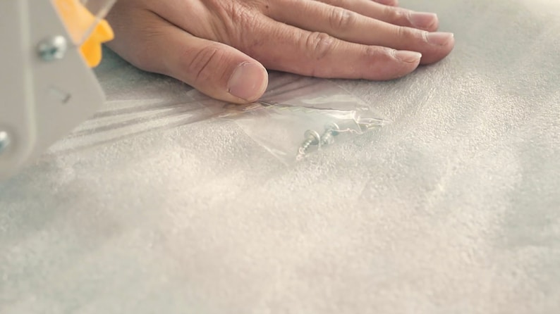 May include: A close-up of a clear plastic bag containing small silver screws. The bag is lying on a white surface.
