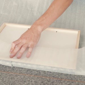 May include: A person is carefully wrapping a wooden frame with white tissue paper. The frame is on a gray surface.