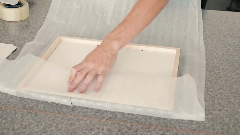 May include: A person is carefully wrapping a wooden frame with white tissue paper. The frame is on a gray surface.