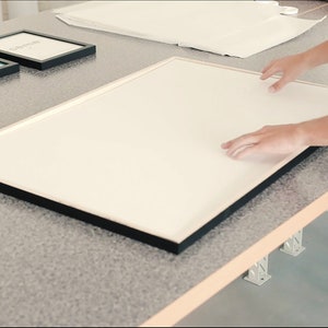 May include: A person places a large, white framed picture on a table. The frame is black and has a simple design. The table is made of wood and has a light brown finish.