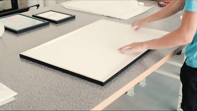 May include: A person places a large, white framed picture on a table. The frame is black and has a simple design. The table is made of wood and has a light brown finish.
