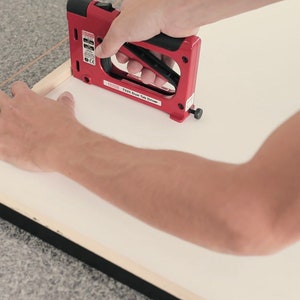 May include: A person using a red electric staple gun to attach a white canvas to a black picture frame.