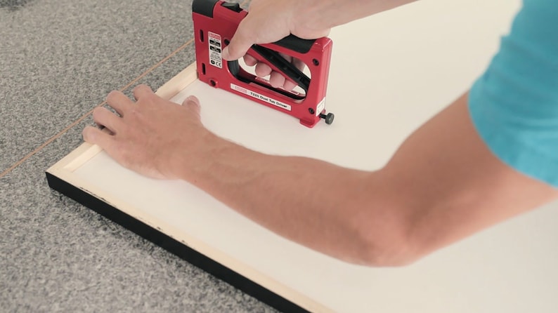 May include: A person using a red electric staple gun to attach a white canvas to a black picture frame.