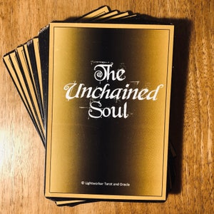 May include: A stack of gold and black tarot cards with the title "The Unchained Soul" and the copyright information "© Lightworker Tarot and Oracle".